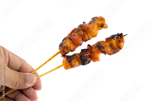 Close-up view of a person's hand holding two sticks of freshly grilled chicken satay on bamboo skewers, isolated on a white background.