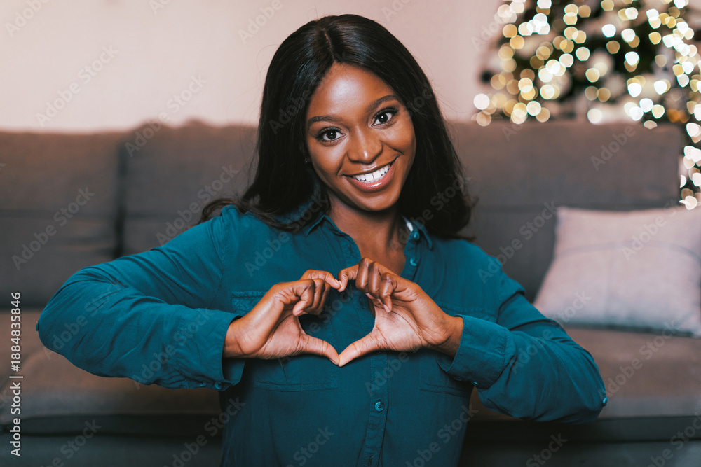 Fototapeta premium A woman with dark hair smiles at the camera while making a heart shape with her hands. She is sitting on a couch in a living room with a Christmas tree decorated with lights behind her.