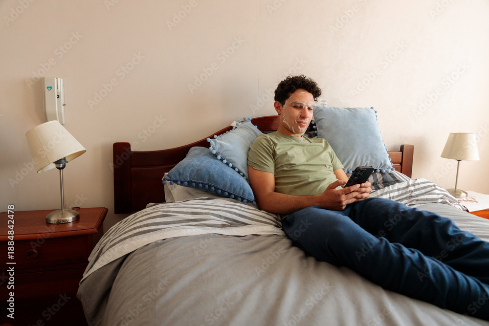 Fototapeta premium Latin man skincare weekend relaxing on bed using smartphone with face mask at home modern masculinity lifestyle