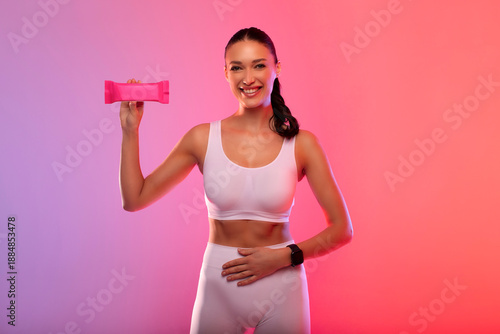 Happy fit woman holding energy protein bar and put hand on belly, having snack after workout, posing over gradient neon background and smiling at camera
