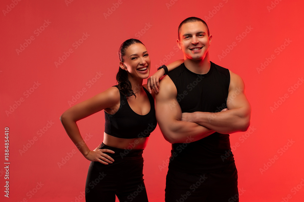 Obraz premium Happy fitness trainer man and woman posing and smiling, both wearing black sportswear, lady putting hand on guy shoulder, standing over red neon background