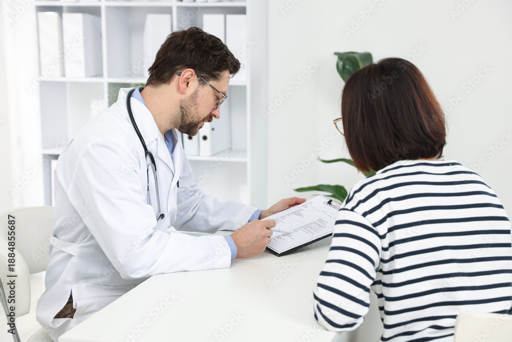 Fototapeta premium Doctor with clipboard consulting patient at white table in clinic