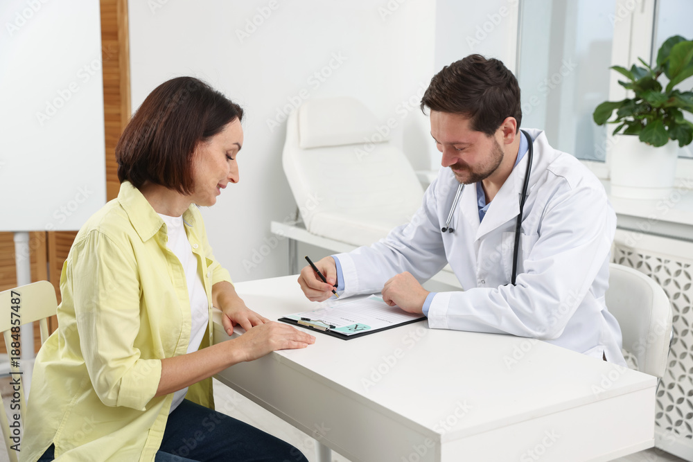 Fototapeta premium Doctor with clipboard consulting patient at white table in clinic