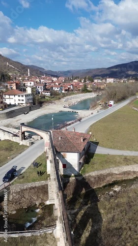 Cinematic vertical drone footage of Tanaro River with ancient Roman bridge and Bagnasco city on the left bank and old mill on the right bank captured on a sunny winter day 3