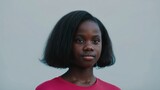 Young black girl with serious expression wearing a red shirt, looking at camera against a plain blue background, portrait