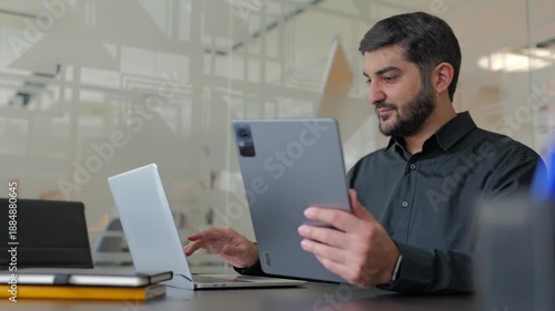 Focused businessman working on laptop and tablet with charts on desk in office.