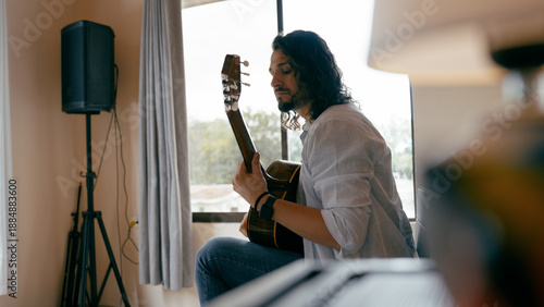 Fotografie Man with long hair sitting by window, concentrating on playing acoustic guitar d