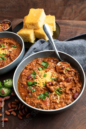 Fotografija A rustic overhead view of homemade beef chili served in bowls and topped with shredded cheddar cheese, fresh herbs, and sliced jalapeños