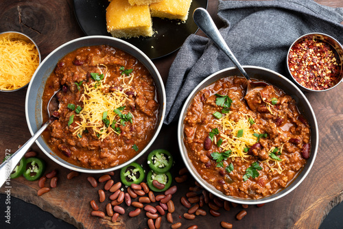 Tablou pe pânză A rustic overhead view of homemade beef chili served in bowls and topped with shredded cheddar cheese, fresh herbs, and sliced jalapeños