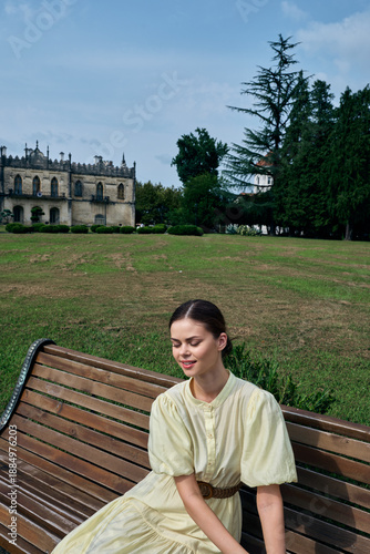 Картината върху платно Woman in a vintage pale dress sits on a wooden bench in a park with a historic c