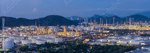 Panoramic night view of a large oil refinery and petrochemical plant located near mountains. Industrial energy estate landscape with illuminated storage tanks and distillation towers at twilight.