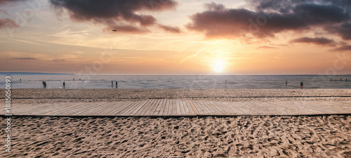 Sandy beach with a wooden boardwalk leading to calm ocean waters, people relaxing and strolling along the shore at a vibrant, colorful sunset with mirrored reflections on the sea