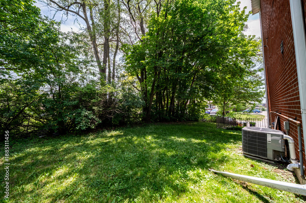 Obraz premium Lush green lawn and mature trees fill a residential backyard on a bright day, featuring a central air conditioning unit positioned against a brick wall of a suburban home