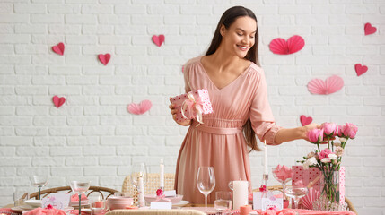 Young woman with gift and roses near festive table setting in dining room. Valentine's Day celebration