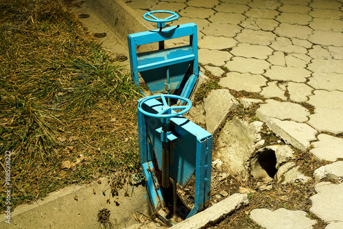 Принти на полотні A close-up shot of a blue metal water control gate, or sluice gate, installed in a concrete drainage channel next to a grassy area and paved sidewalk at night