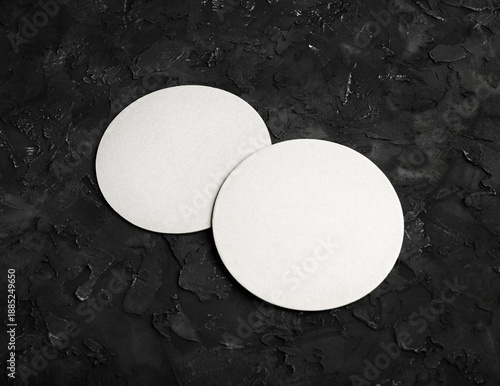 Two blank white beer coasters on on black textured surface.