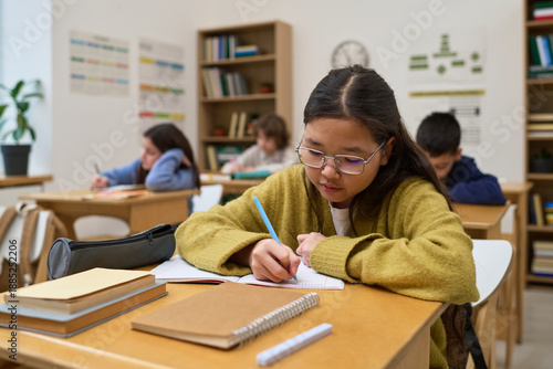 Photos Asian preteen girl sitting at desk writing in notebook in classroom with other c