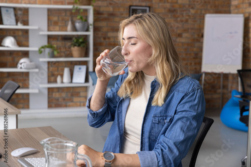 Female office worker sitting at desk drinking water from clear glass near keys wearing denim shirt