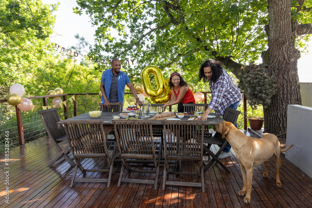 Fototapeta premium Diverse friends arranging party setup on wooden deck with dining table gold '0' balloon, dog