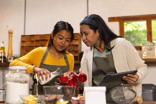 Foto Diverse mother and daughter pouring milk into red measuring cup in home kitchen,