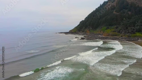 An aerial, wide shot view of the dramatic and rugged coastline at Cape Perpetua, Oregon. Large waves crash onto the expansive, rocky beach and a steep, forested bluff rises from the shore under an ove