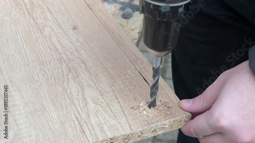 A person drills through an angled wooden piece with a cordless electric drill on a workbench outdoors