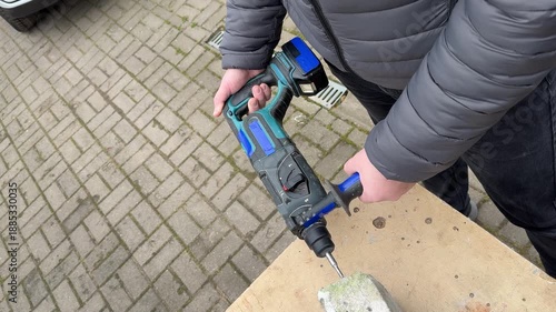 A person drills a vertical hole into the edge of a concrete block with a cordless hammer drill using a two hand grip, mid shot.