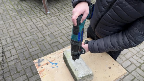 A person drills a hole with a cordless hammer drill into a concrete block near the edge while standing outdoors, angled view.