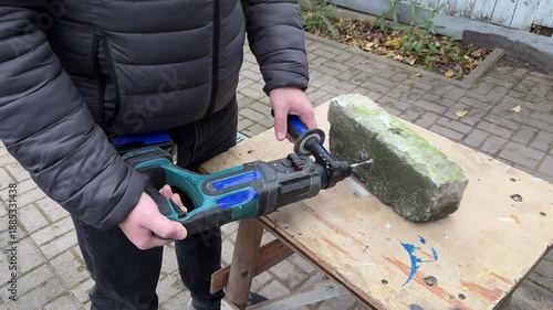 
A person drills a hole with a cordless hammer drill into the edge of a concrete block on a workbench outdoors, close up.