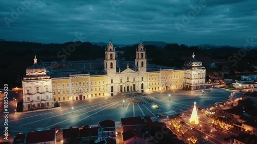 Aerial drone approach toward Mafra National Palace at dusk, starting from a slightly lateral and distant angle, slowly moving forward to a closer view of the illuminated facade under blue hour sky.