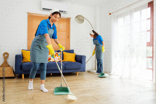 Two uniformed cleaning professionals sweep and maintain a modern living space with wooden floors and a blue sofa, showcasing efficient, reliable facility care in a professional service setting.