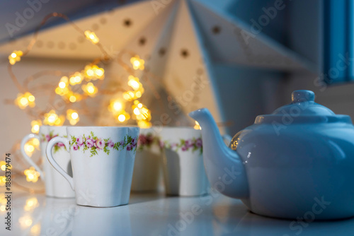 White porcelain teapot and cups with floral patterns against a backdrop of golden star lights. Winter time