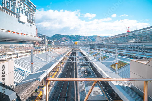 Modern Japanese Train Station with Commuter Train and Urban Mountain Background under Blue Sky High-angle view of a modern Japanese railway station with a commuter train in motion, platform roofs,