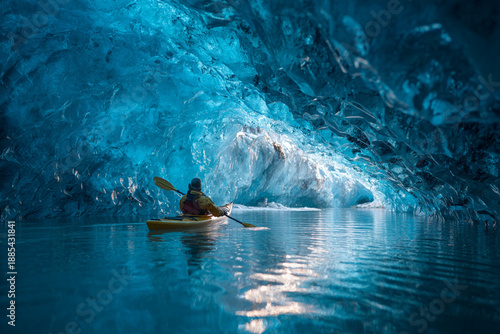 Adventurous kayaker explores the breathtaking depths of a brilliant blue glacial ice cave paddling through calm reflective water toward bright opening.