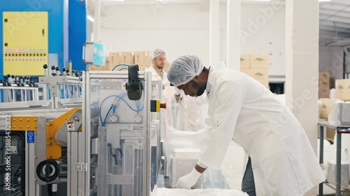 African American male lab worker wearing protective coat and hairnet packaging aluminum trays on production line. Focused man working at automated food container factory. Industrial setting indoors.