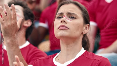 Worried Caucasian female football fan with red and white face paint clasping hands watching tense match. Woman in red jersey reacting emotionally in stadium crowd. Outdoors. Close up.