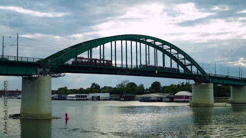 Urban landscape of an old iron bridge above a river with anchored restaurant rafts and a dramatic sky.