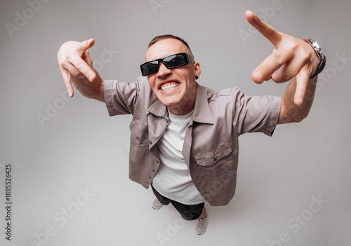 Studio portrait of a young man in gray shirt and sunglasses making expressive rapper hand gestures and facial expressions. Fisheye top-angle lens creates strong body distortion on gray background.