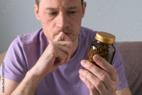 Фотография Skeptical man holds jar of capsules and thinks about supplement choice, decides whether to take pills or skip them for safety