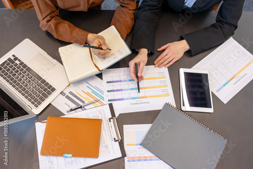 Business professionals collaborating at a desk, reviewing financial reports and spreadsheets while taking notes and using a laptop and tablet, working together on a strategic plan