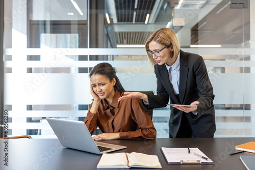 Strict female boss scolding a pressured female employee looking upset and holding her head, experiencing bullying and workplace conflict in a modern office environment