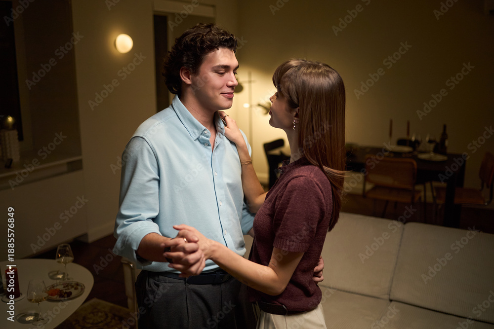 Fototapeta premium Caucasian young adult man and Caucasian young adult woman dancing together in living room, holding hands and looking at each other, sharing intimate moment indoors
