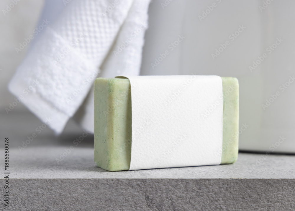 Fototapeta premium Green soap bar with blank label near white bath towels and basin on grey stone, mockup