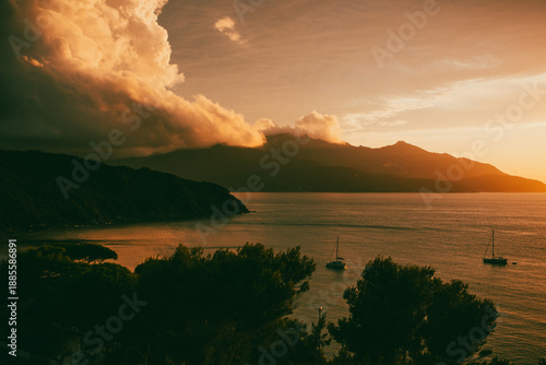 Serene coastal sunset with sailboats anchored in calm waters, under a dramatic cloud-filled sky. Elba Italy