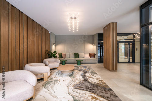 Modern lobby with wooden wall panels, plush armchairs, long sofa with cushions, abstract rug, wooden coffee table, stylish pendant lighting, green plant accents.