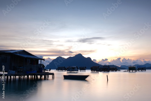 Fotografía Wooden boat floating near stilt houses at sunrise