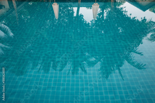 Billede på lærred A refreshing view of blue tiled swimming pool water surface reflecting silhouettes of coconut palm trees and white umbrellas at a tropical resort during the day