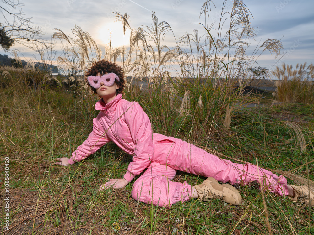 Fototapeta premium Person in pink jumpsuit and mask lying on a grassy field at sunset, playful outdoor cosplay look with dramatic pose and natural background, rich color and texture