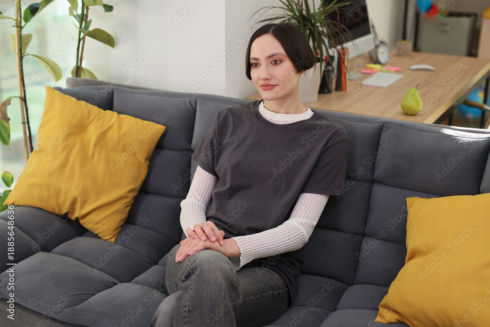 Obraz premium In a serene living room, a young woman sits comfortably on a modern couch, lost in thought as soft sunlight filters through the windows, illuminating her surroundings.