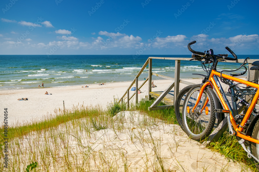Fototapeta premium Scenic view of Curonian Spit beach with orange bicycles parked on grassy dunes near wooden stairs overlooking Baltic Sea coastline in summer